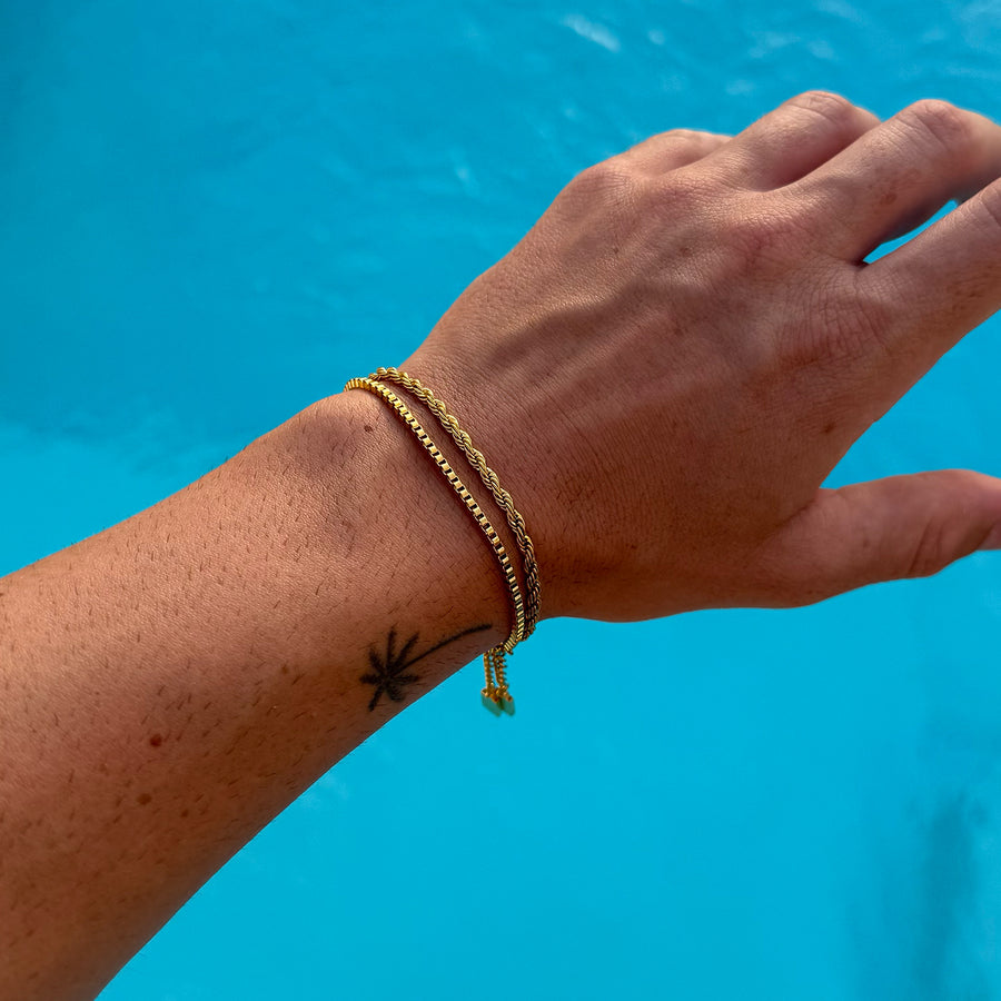 Gold snake chain bracelet by FRNE worn on wrist against blue water background