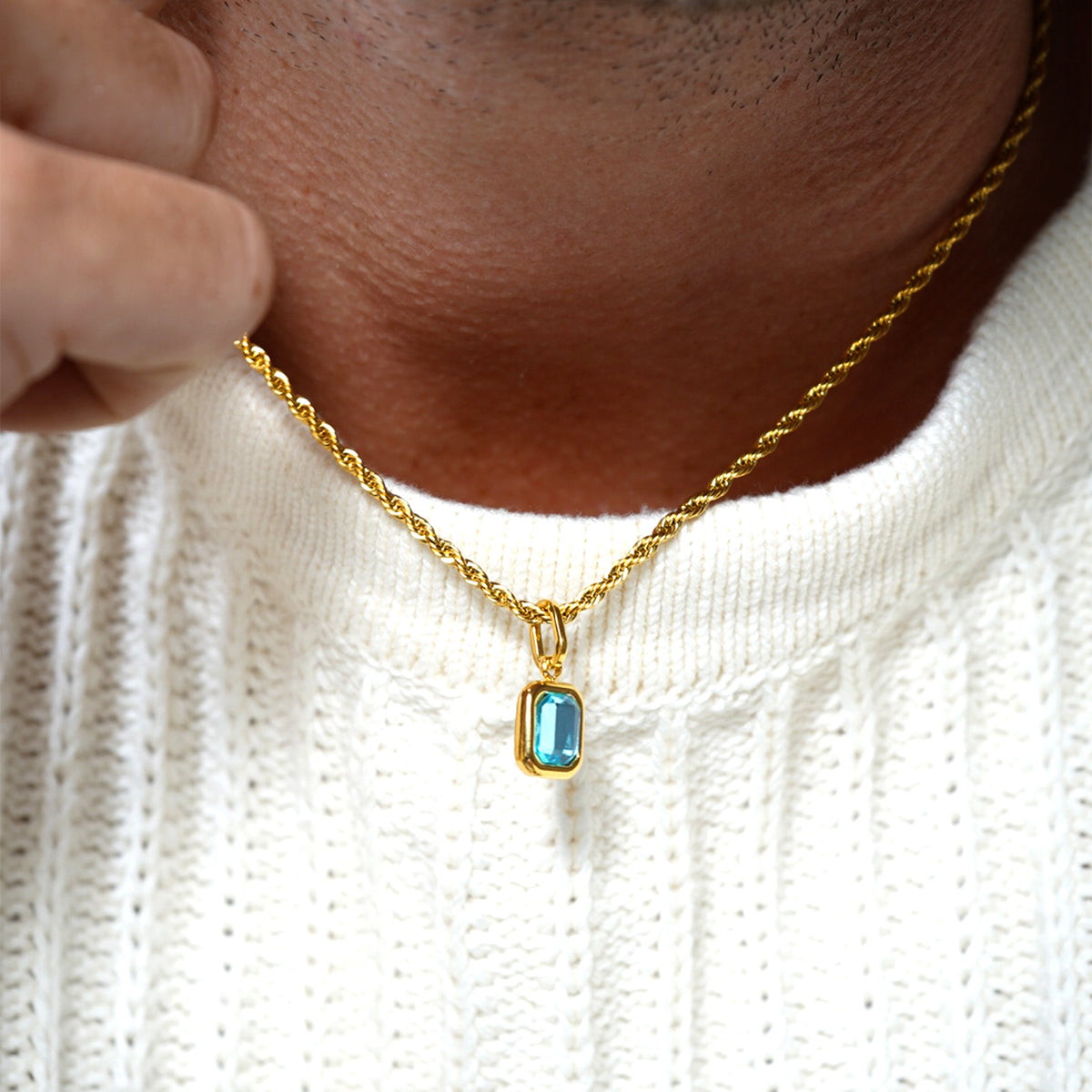 Close-up of FRNE glacier blue gemstone gold pendant necklace worn by a person, showing a rectangular light blue stone set in a gold frame on a twisted gold chain against a textured white sweater