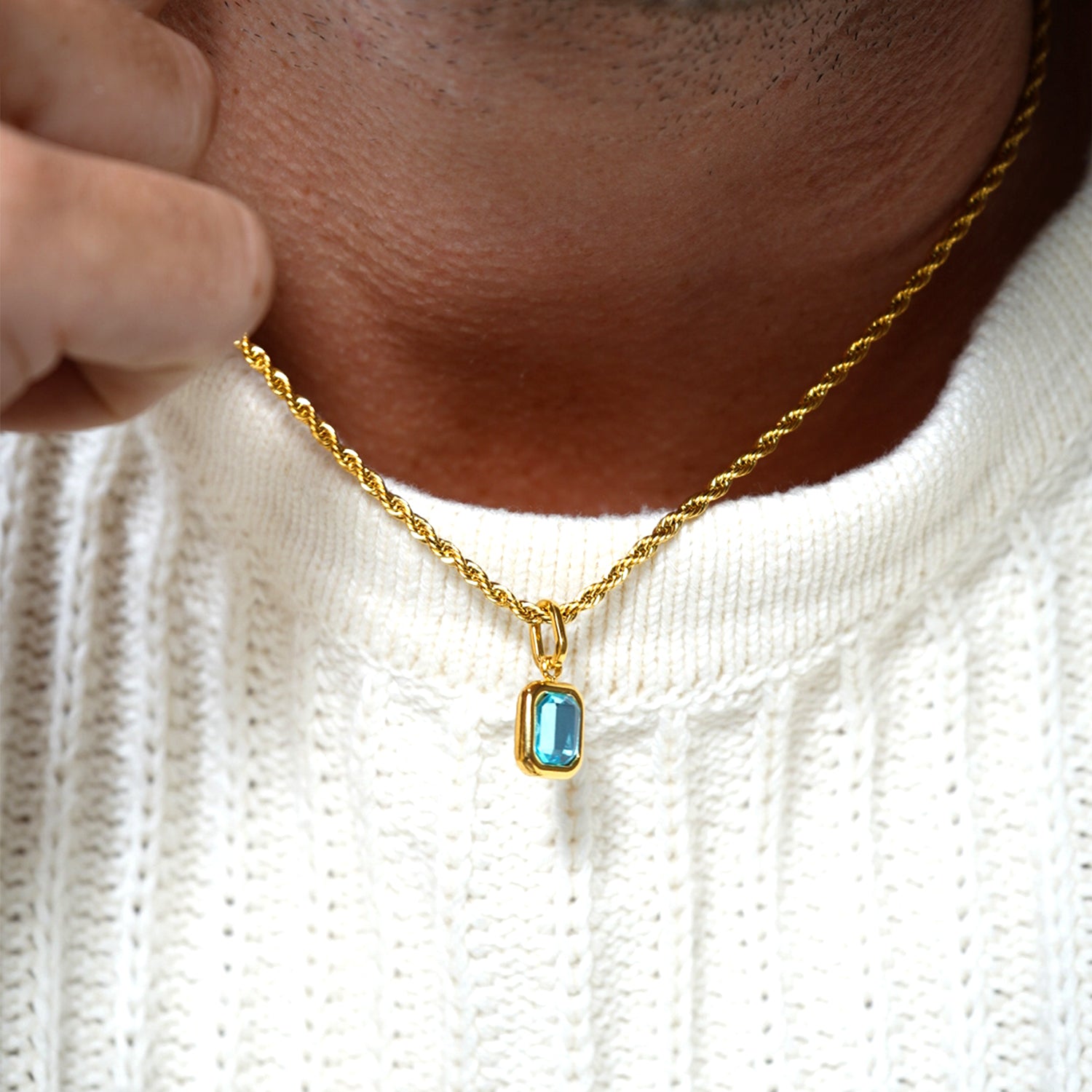 Close-up of FRNE glacier blue gemstone gold pendant necklace worn by a person, showing a rectangular light blue stone set in a gold frame on a twisted gold chain against a textured white sweater