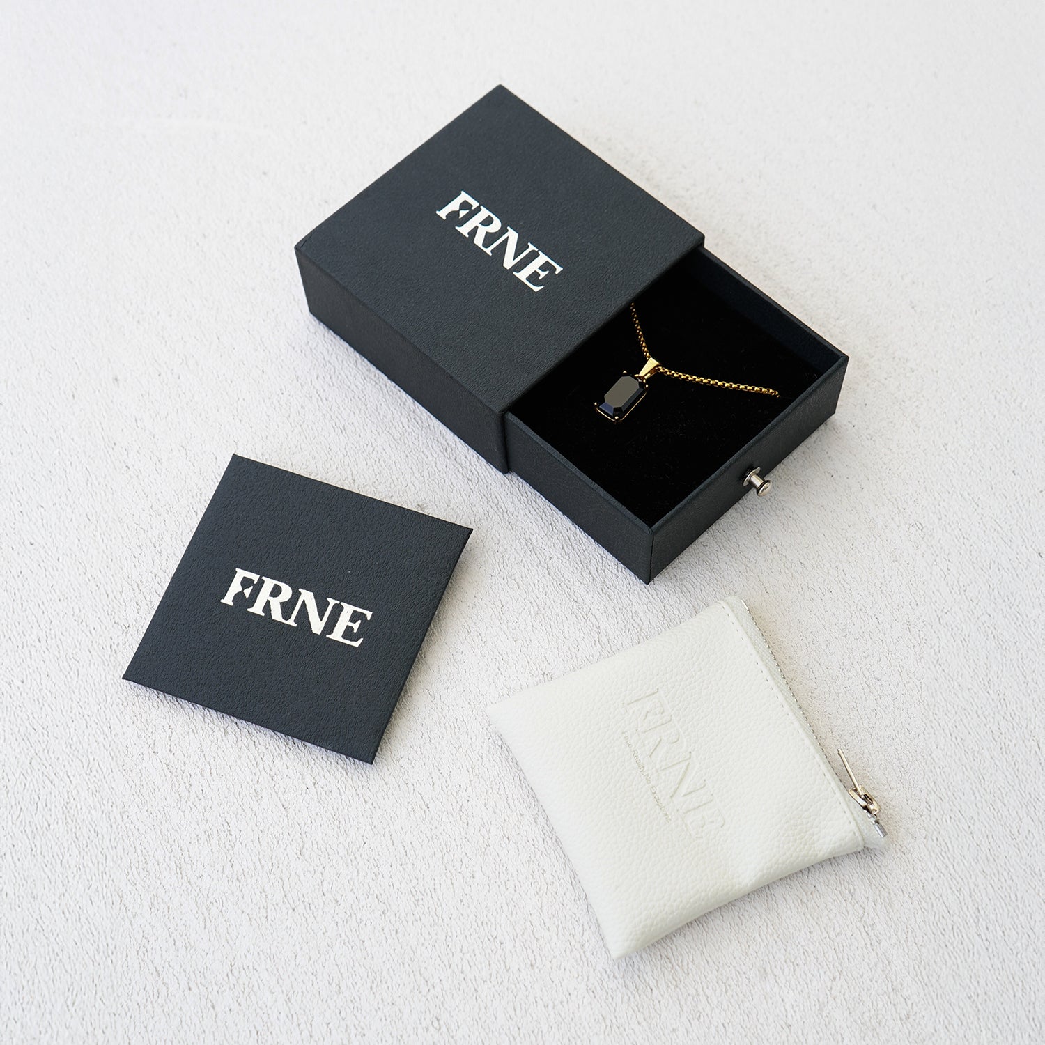FRNE jewellery set with gold necklace in a black box alongside branded card and white pouch on a clean background