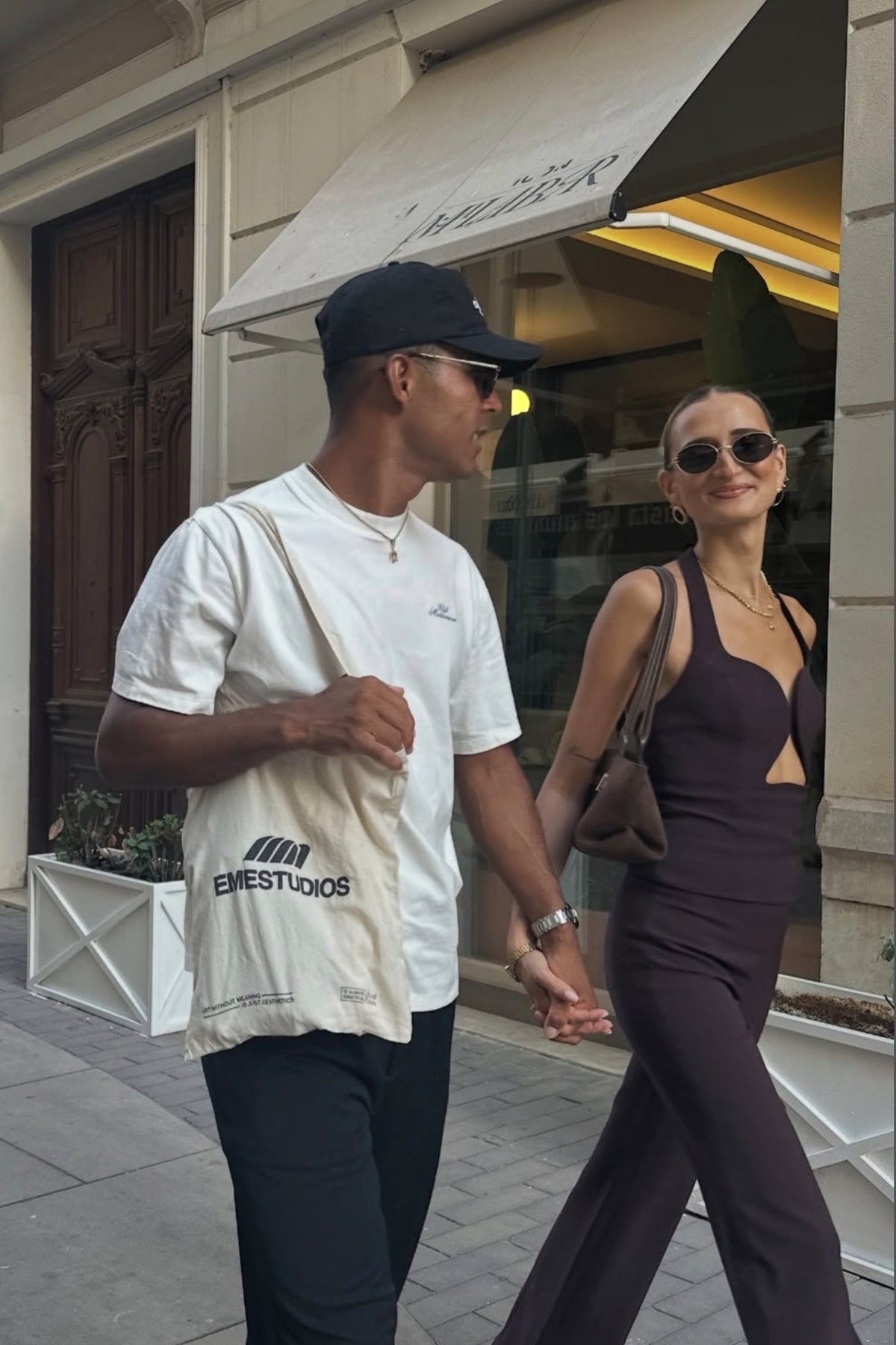 Stylish couple walking hand in hand on a city street, wearing casual outfits and accessories in front of a storefront with an awning